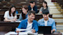 Two students sitting a lecture hall while looking at a laptop, with three other students seated behind them.
