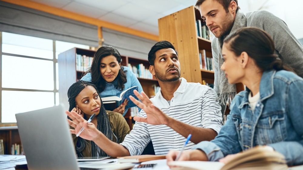 A group of international graduate students have a discussion around a table at a library over open books and notebooks 