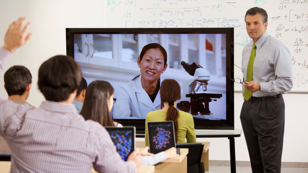 There are many types of professors at US universities. In this photo, a faculty lecturer stands in front of a whiteboard, next to a television broadcasting a guest speaker, in front of a class of international students. 