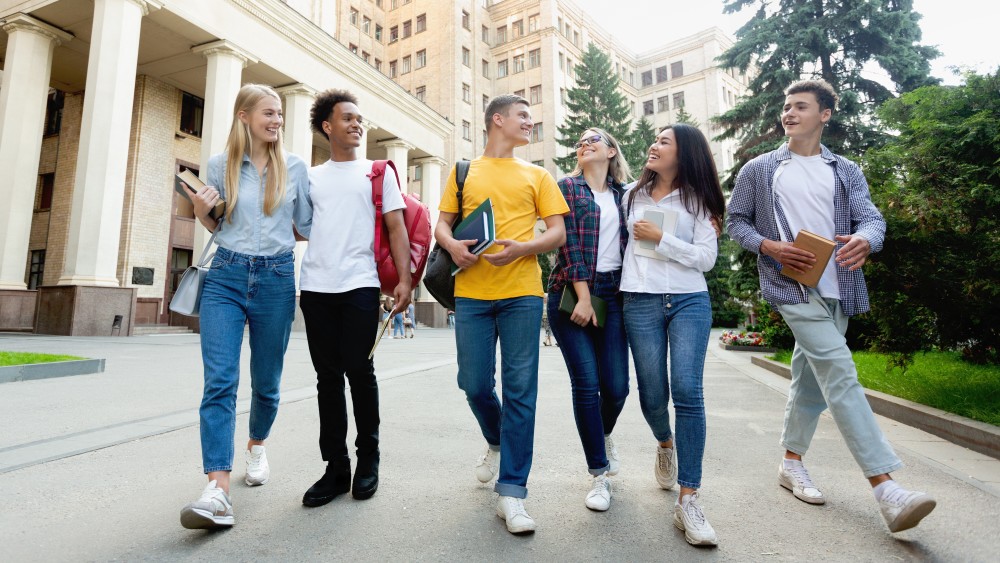 A group of international students holding textbooks walk and laugh together on a city campus