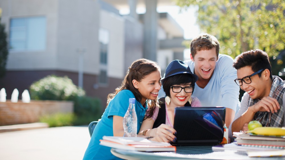 Two female and two male international students at a US university campus sit outside at a table in front of a tree and look at a laptop