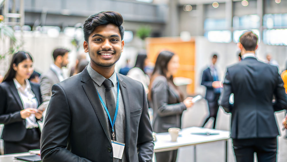 A male international student from South Asia wearing a suit stands in an exhibition hall for a professional networking event at his US university.