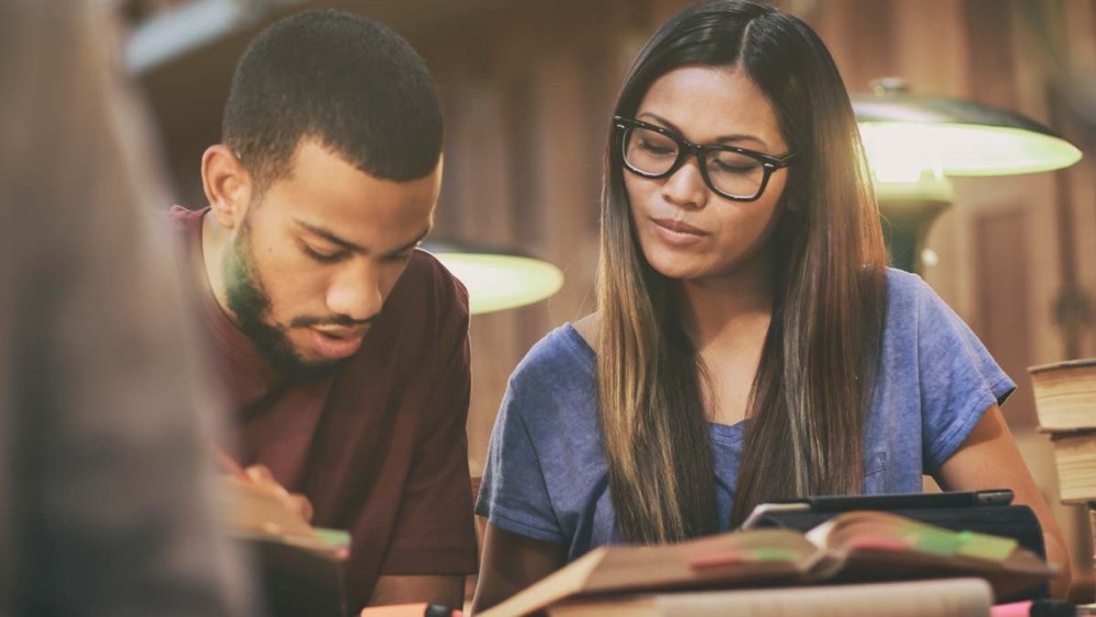 A male international student and female international student study side by side in front of a book in a university library. 