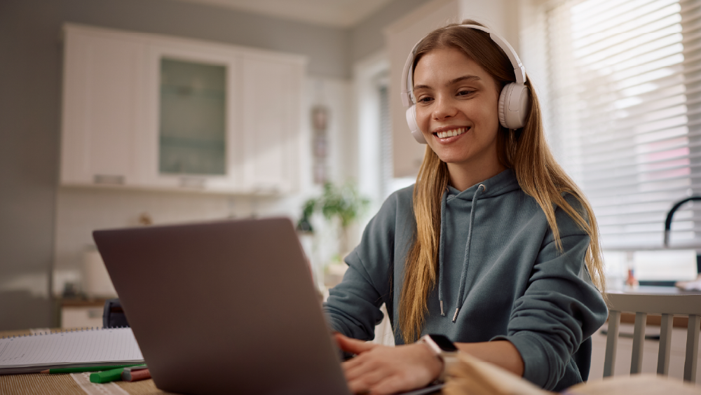 A female international student wearing headphones sits at a table in front of a laptop for her online class.