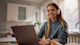 A female international student wearing headphones sits at a table in front of a laptop for her online class.