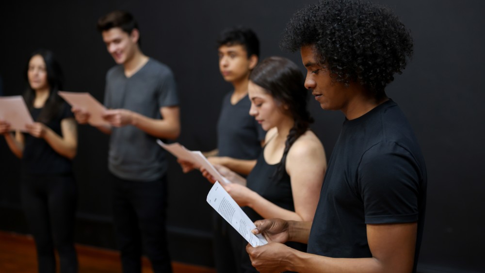 What are electives? Electives are courses at US universities that count toward graduation but may not be directly related to a student's major or degree program. In this image, a group of three international students taking an elective course at a US university stand in a row and study scripts for a theater exercise. 