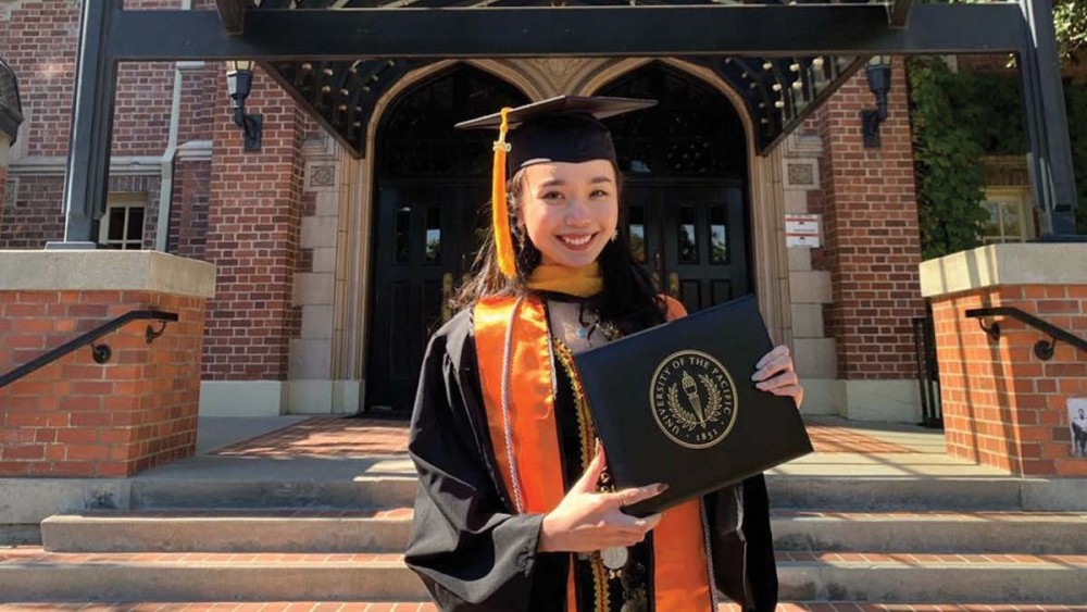 Jacqueline, an international student from Malaysia at the University of the Pacific, stands in front of a brick building on campus in her graduation cap and gown and holds up her diploma
