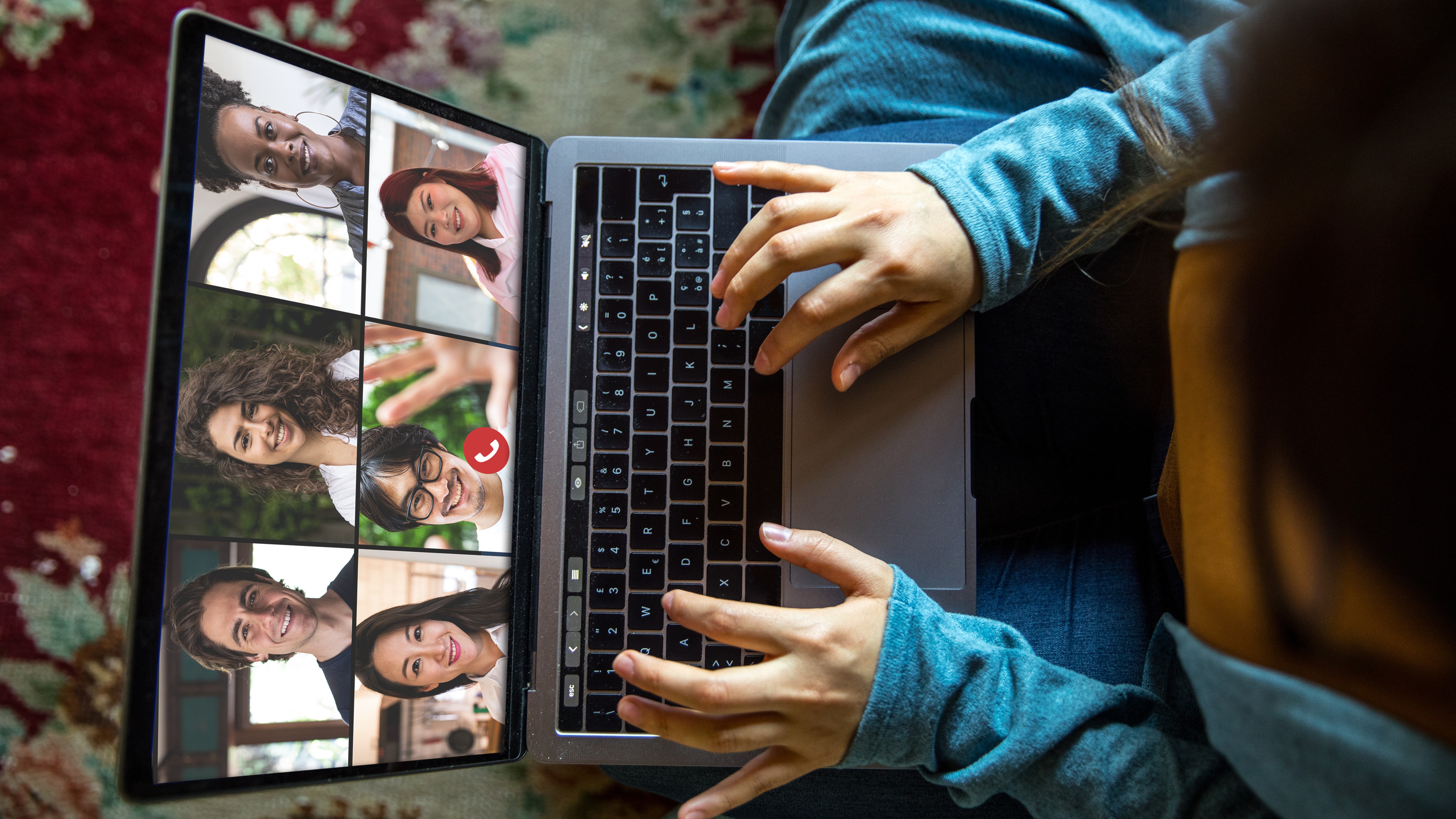 An international student types at a laptop during a videoconference meeting with six attendees on screen