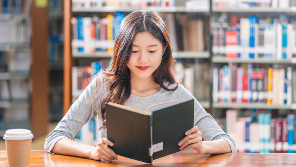 A female Asian international student sits in her US university library and reads an English book with a cup of coffee.