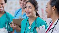 A female international student in medical school in the US wears scrubs and a stethoscope and smiles while holding a clipboard