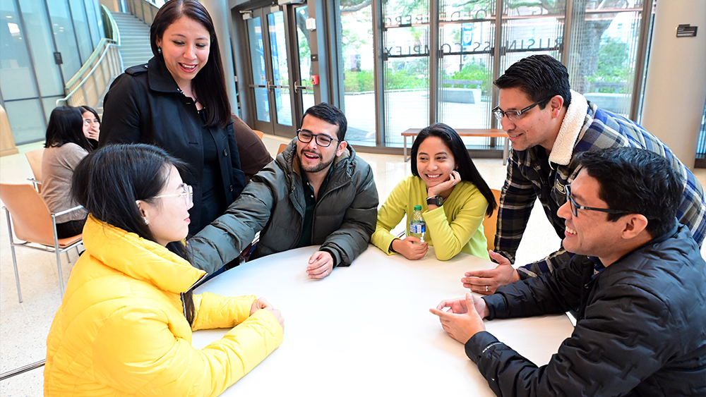 A group of international students at the Freeman School of Business at Tulane University sit around a table in a sun-filled atrium and discuss their master's degree in business curriculum. 