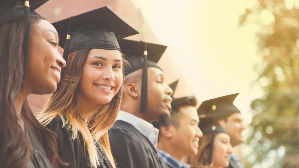 After earning their higher ed degree from the US education system, six students wearing graduation caps and gowns stand while a woman in the middle smiles at the camera.