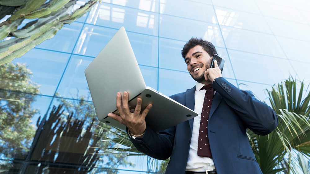 An international student now working in Dubai stands outside a skyscraper wearing a suit and tie, holding a laptop, and talking on a cell phone