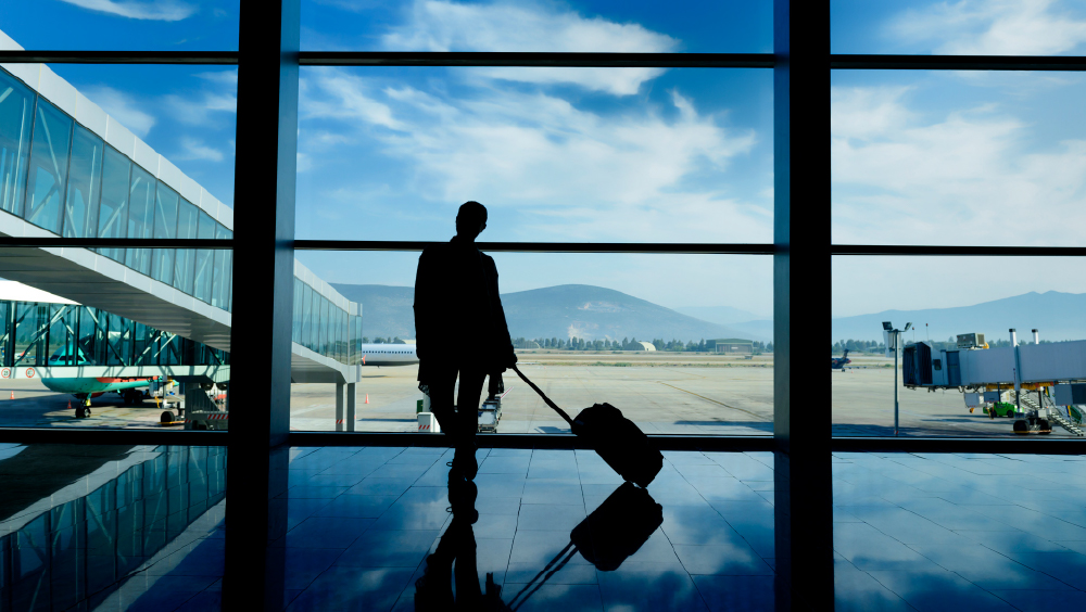 An international student with a suitcase stands in silhouette in front of a wall of windows overlooking an airport tarmac.