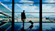 An international student with a suitcase stands in silhouette in front of a wall of windows overlooking an airport tarmac.