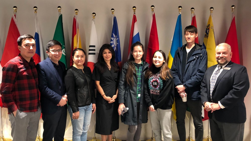 A group of five international college students and managing director Sharmeen Ahsan-Bracciale stand in front of a row of international flags at American University