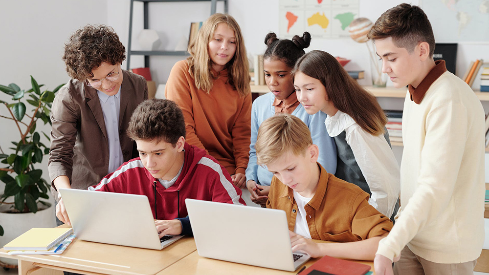 A group of 6 young students with their international student teacher instructor gather around a desk in a classroom and review ed tech software on two laptops.