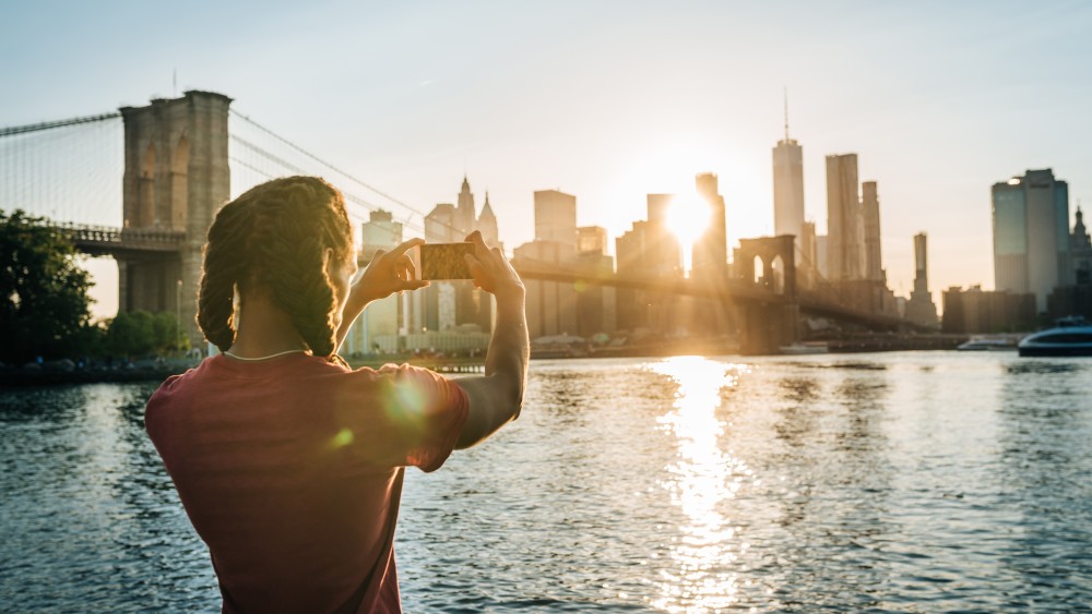 An international student stands in front of a river against the Brooklyn Bridge and NYC skyline at sunset taking a photograph. 