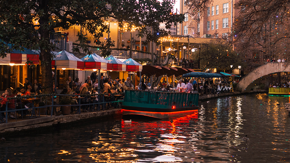 A bottom-lit riverboat passes restaurants with outdoor patios in early evening along the San Antonio River Walk.
