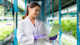 A female international student wearing a lab coat, working toward a food technology specialization for her US university degree, stands with a laptop in a greenhouse near rows of leafy green plants.