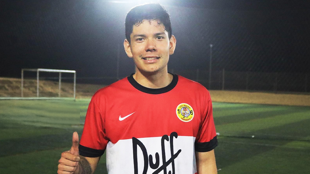 Rodrigo, an international student from Peru at Heriot-Watt University, wears a Duff football jersey and gives a thumbs-up while standing on a football pitch.