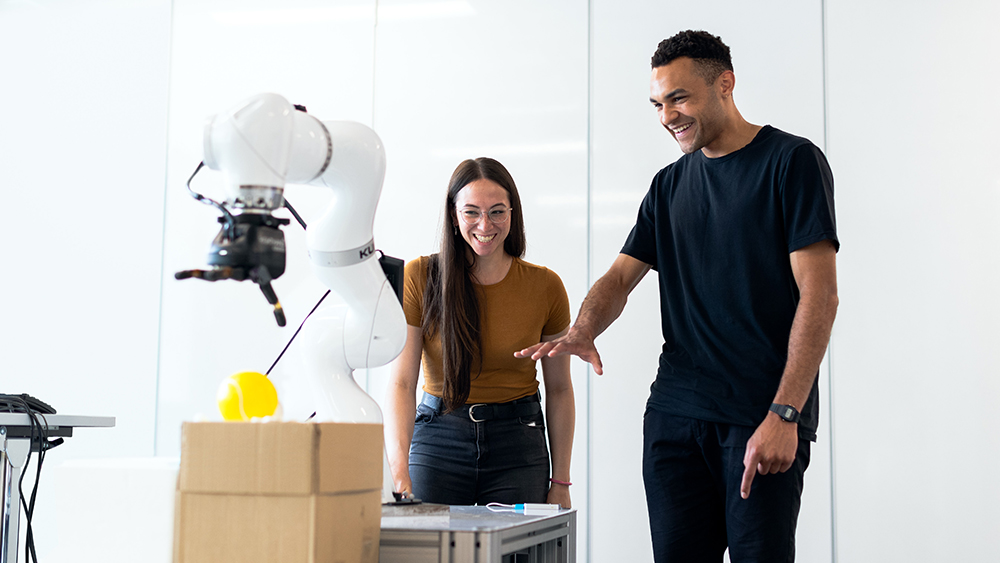A female international student and a male international student stand in an engineering lab and work on a prototype