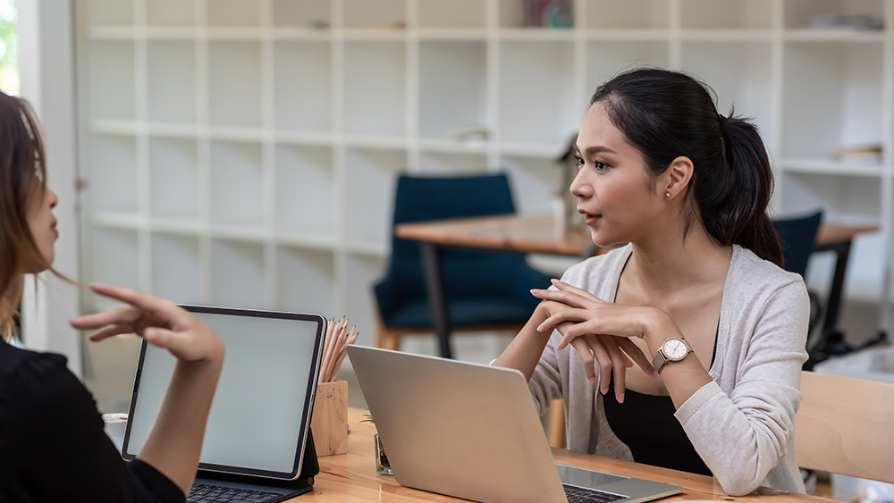 A female international student sits at a table in front of an open laptop and speaks with her education advisor about getting an academic transcript to submit with her US university application.