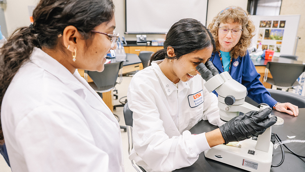 Two Mexican female PhD candidates at The University of Texas at San Antonio work with a microscope in a UTSA lab while their professor looks on.  
