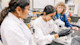Two Mexican female PhD candidates at The University of Texas at San Antonio work with a microscope in a UTSA lab while their professor looks on.  
