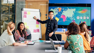 A group of international students sit around a conference table while one student makes a communication skills presentation at a easel in front of a world map