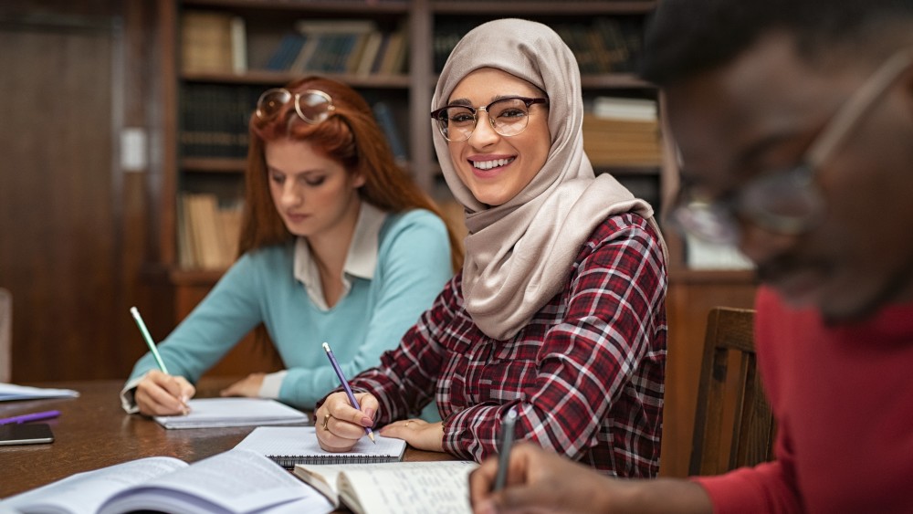 A smiling Pakistani student in a hijab sitting next to an American student both taking notes in a college classroom.