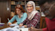 A smiling Pakistani student in a hijab sitting next to an American student both taking notes in a college classroom.