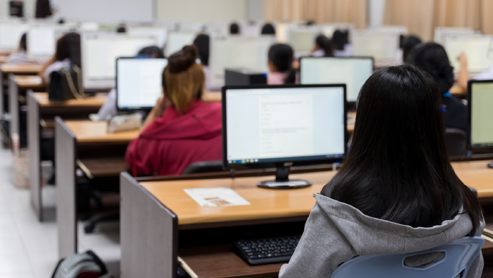 Students working at desktop computers in a classroom computer lab, viewed from behind.