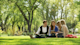A group of four international students sits on a lawn under a grove of trees on the quad at their US university campus during the spring semester