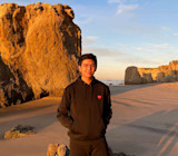 Damian, an international student from Indonesia at American Collegiate, Los Angeles, stands on the beach in front of rock boulders at sunset