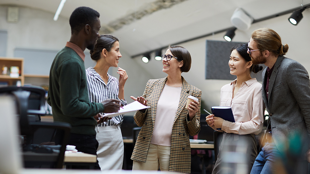 A group of international students working at an internship at a startup stand in a conference room and discuss their latest project.
