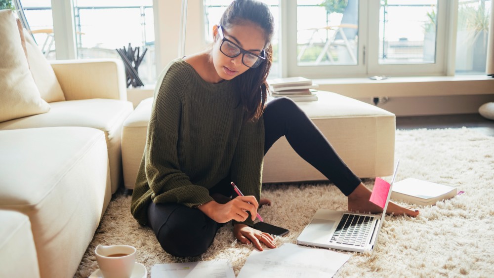 A female international student from Asia wears glasses and sits on the floor in front of a laptop and notebook as she works on a paper before the deadline