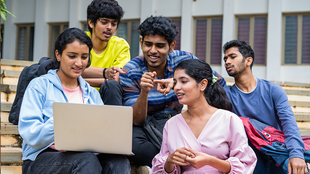 A group of international students from Bangladesh sit outside at their US university and work on a group project together on one laptop.