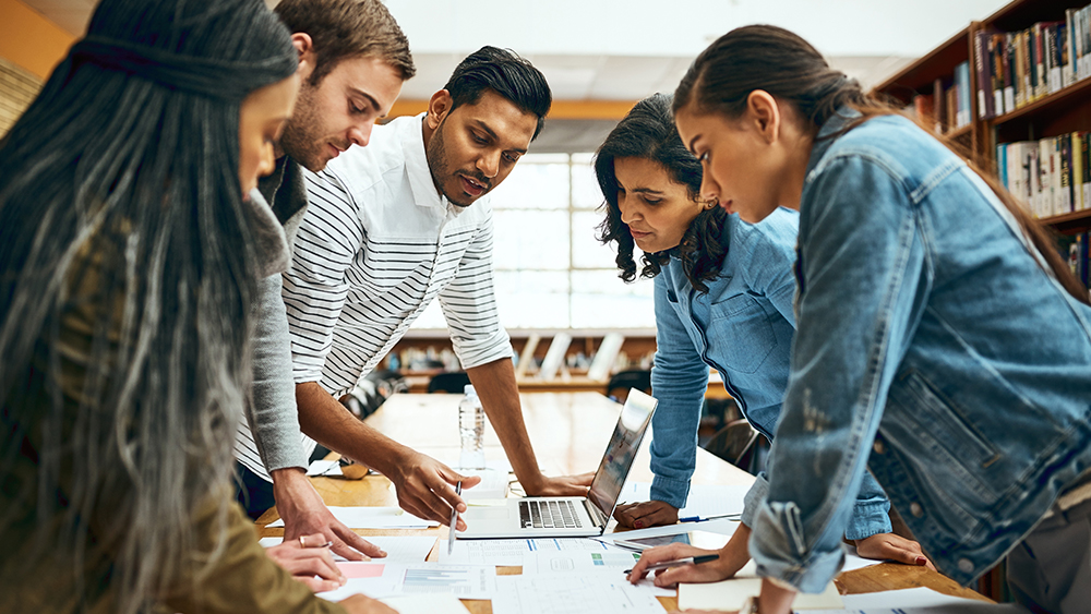A group of international students in a pathway program at a US university stand around a classroom table with an open laptop and papers as their professor goes over an assignment.