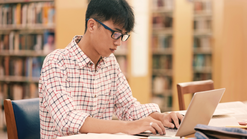 An Asian male international student sits at his laptop in his US university library and works on his dissertation.