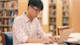 An Asian male international student sits at his laptop in his US university library and works on his dissertation.
