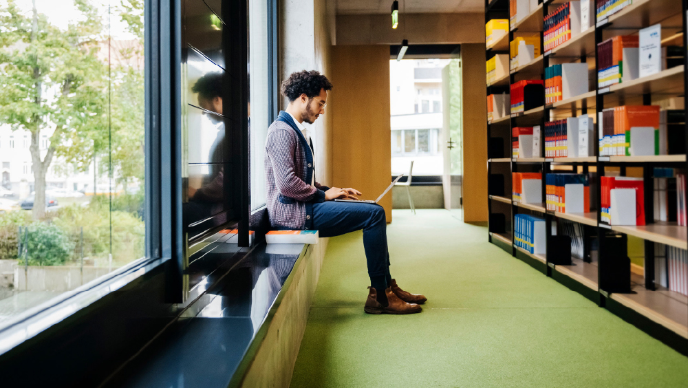 A male international student sits on a windowsill across from bookshelves at his US university library and works on a laptop.