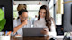 Two female international students sit side by side in front of a laptop