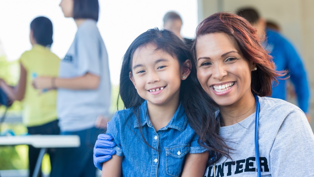 A female college student wearing a "volunteer" t shirt with her arm around a young girl, and both are smiling at the camera.