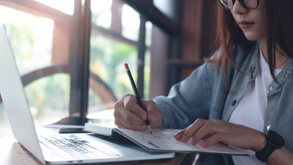 A female international student from Asia sits at her desk with her laptop and day planner and fills out appointments in her US school year calendar.