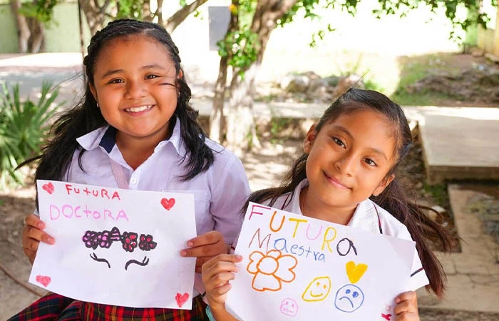 Two young Hispanic girls smile at the camera. They each hold up a piece of paper with colorful drawings.