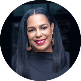 A black woman with long, dark straight hair wears a black blouse and large gold earrings as she smiles at the camera.