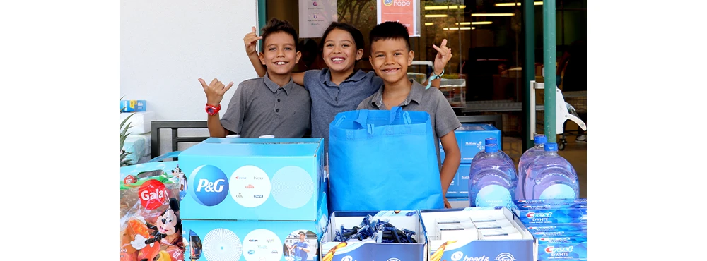Three young school aged children in gray collared shows doing the Hawaiian Shaka hand gesture, standing behind a table of P&G disaster relief kit boxes, bags and products.