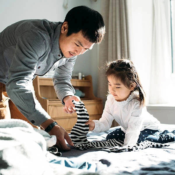 A father leans over his daughter. He holds a garment in his hand as he teaches her how to fold it.