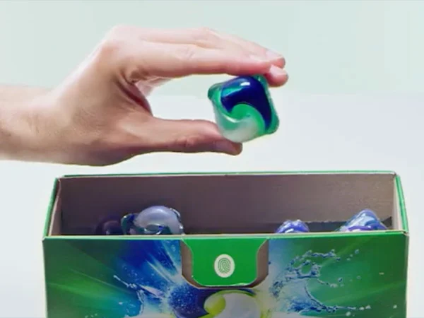 A hand holds a multi-colored laundry detergent pod. Underneath the hand is a blue and green box with more detergent pods.